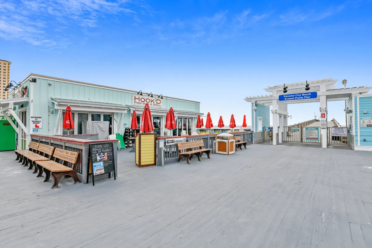 A charming restaurant facade with bright red umbrellas shading wooden benches and tables, inviting guests to dine or socialize outdoors. The welcoming entrance and casual atmosphere suggest a lively spot for evening gatherings close to the beach.