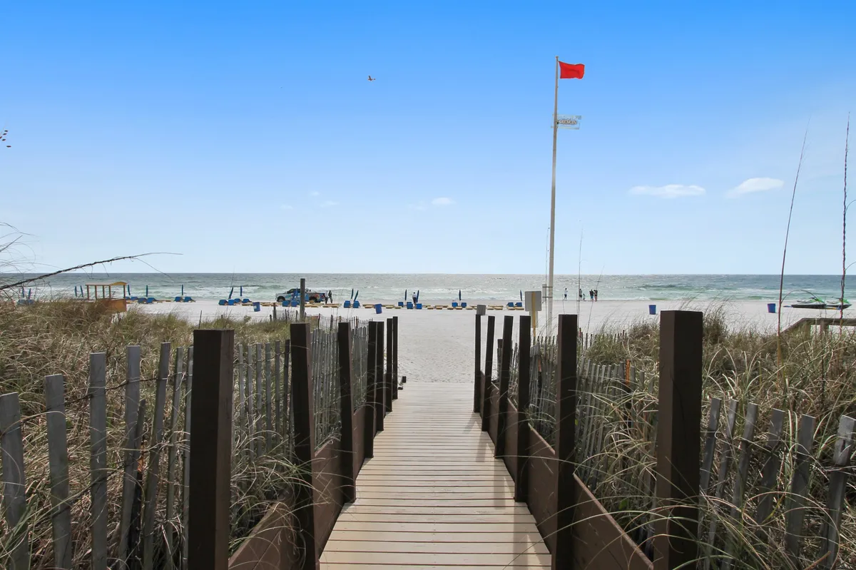 A sunlit wooden boardwalk leads to a pristine stretch of beach, with gentle ocean waves in the background and a scattering of beach chairs ready for relaxation. A red flag flutters in the sea breeze, and the scene is framed by coastal vegetation, evoking a sense of peaceful beachside escape.