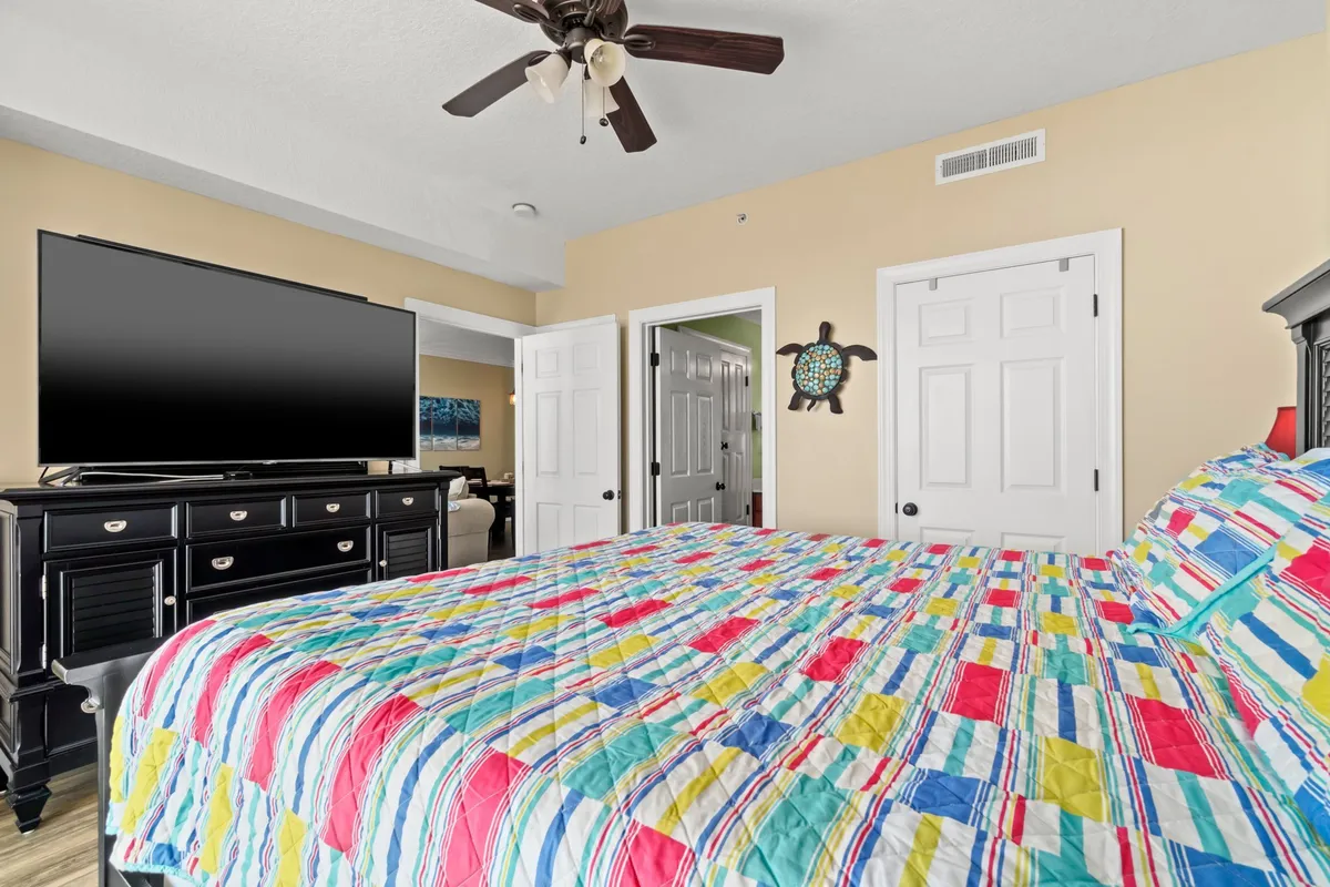 The photo showcases a bedroom with a vibrant, quilted bedspread, a large TV, a modern black dresser, and playful decorative wall art. The space feels both cheerful and restful, with thoughtful details.