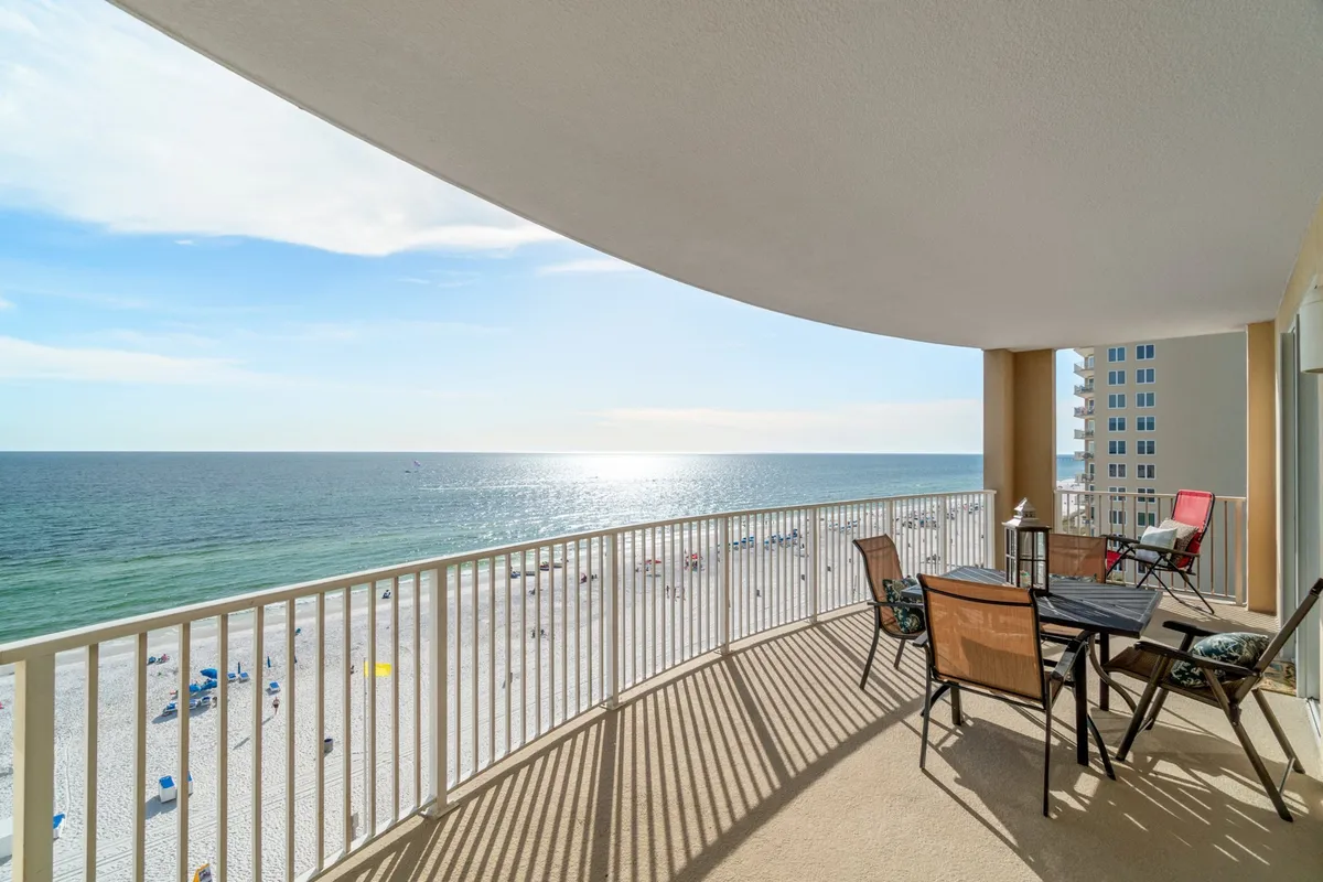 A serene exterior shot of the balcony, outfitted with inviting furniture, overlooking the calm ocean and dotted with colorful beach umbrellas below. The view stretches out to the horizon, promising stunning sunrises and sunsets, while the setup suggests an ideal spot for sipping morning coffee or an evening toast.