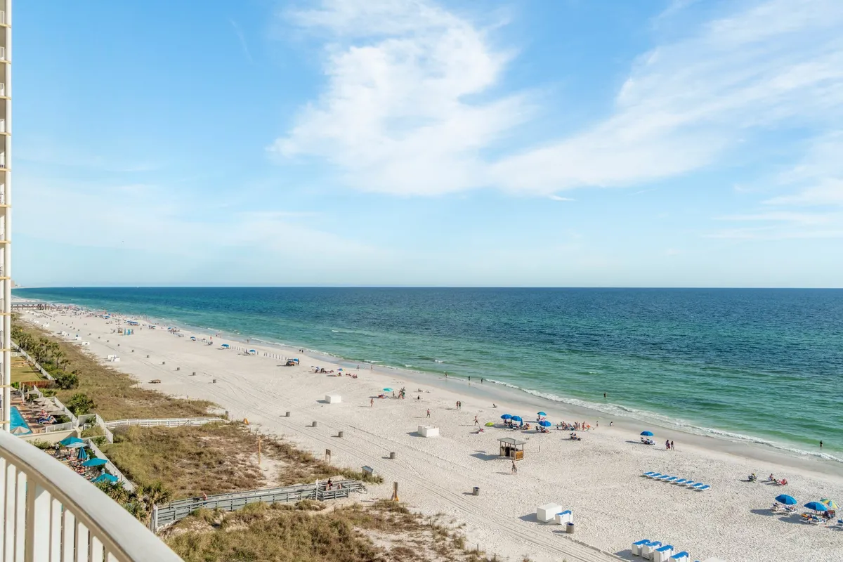 A vibrant beachfront scene shows families and friends scattered along the shore, sunbathing, playing in the sand, and enjoying the Gulf's gentle waves. Colorful umbrellas and beach chairs dot the sandy expanse, while the sparkling ocean stretches to the horizon.