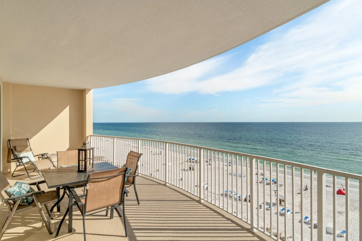 This image captures a tranquil balcony scene overlooking the beach and ocean. The balcony features a sturdy railing, a dining table set for outdoor meals, and comfortable seating, all set against an expansive backdrop of sand, sea, and sky. The soft lighting and gentle clouds create an inviting atmosphere for both daytime relaxation and sunset moments.