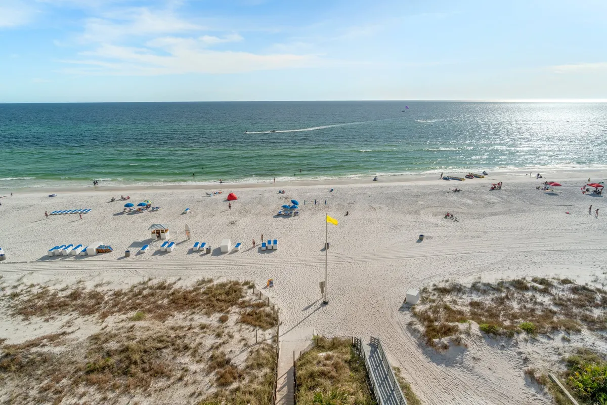 A wide-angle view of the sandy beachfront, dotted with lounge chairs and umbrellas, as people enjoy sunbathing, playing, and strolling along the shore. The vibrant energy of the beach is evident, and the ocean horizon beckons in the distance.