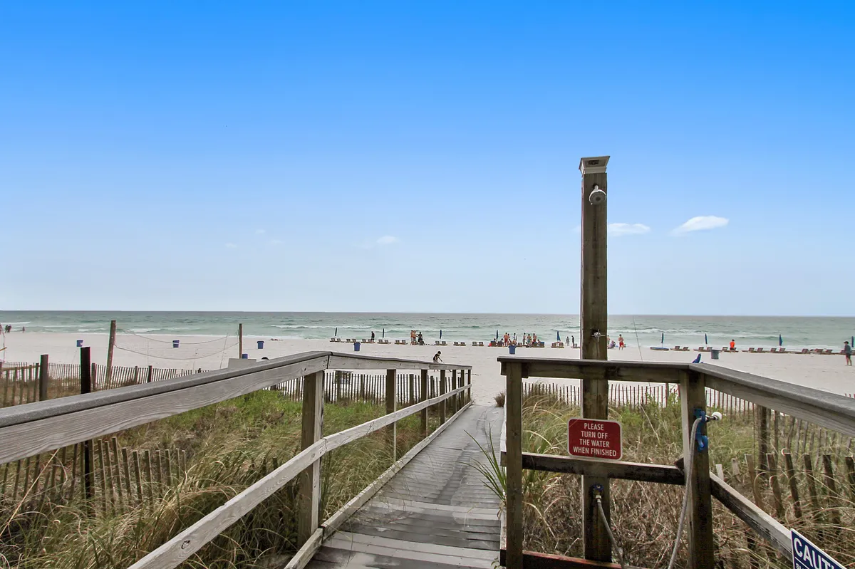 The image features a wooden walkway stretching over sand dunes toward the beach, with the ocean and a few beachgoers in the distance. The scene is serene and highlights the natural beauty and accessibility of the coastline.