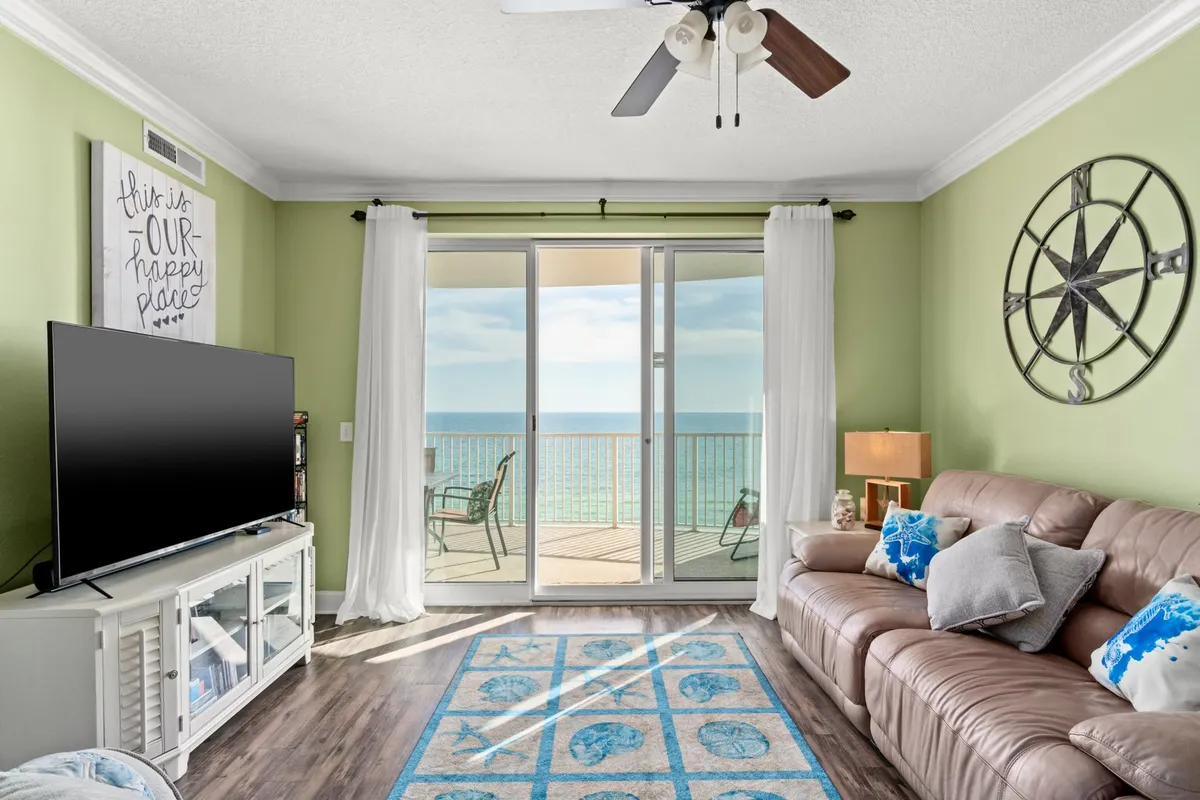 A stylish living room featuring a plush leather sofa, a colorful area rug, a TV unit, and a large window with sweeping ocean views. The room feels both tranquil and cheerful, perfect for relaxing or gathering for a movie night.