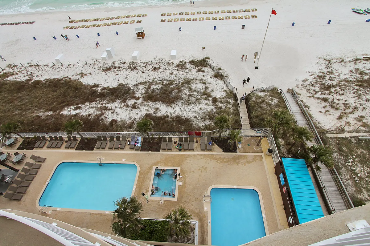 An enticing view of the resort-style amenities, with two sparkling swimming pools, a bubbling hot tub, and comfortable lounge chairs set beside the sandy beach. The ocean is visible just beyond, linking the pool area directly to the Gulf. The scene exudes a sense of leisure and luxury.