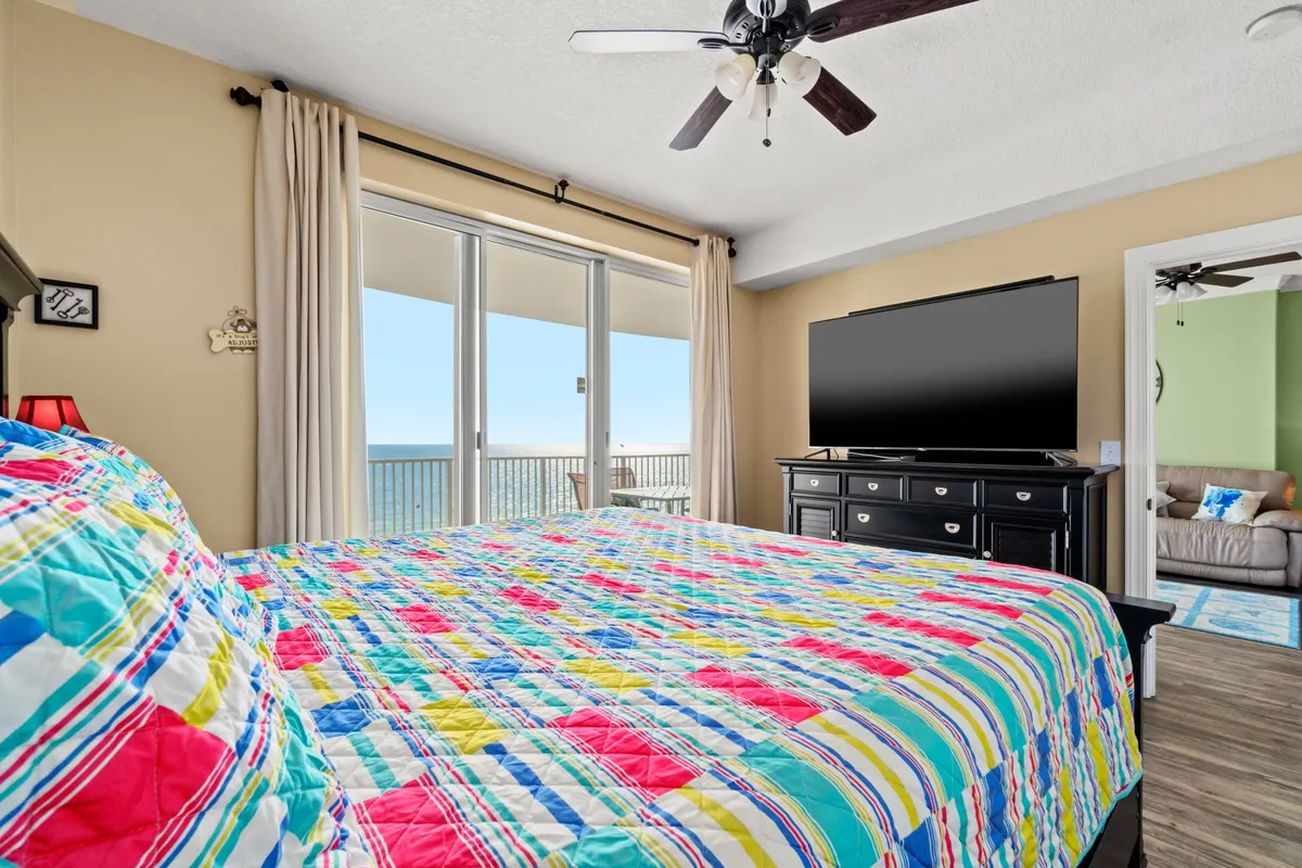 This image features a comfortable bedroom with a colorful quilt on the bed, a large window offering an ocean view, and a television on a dresser. The space is filled with natural light, and the décor feels cheerful and inviting.