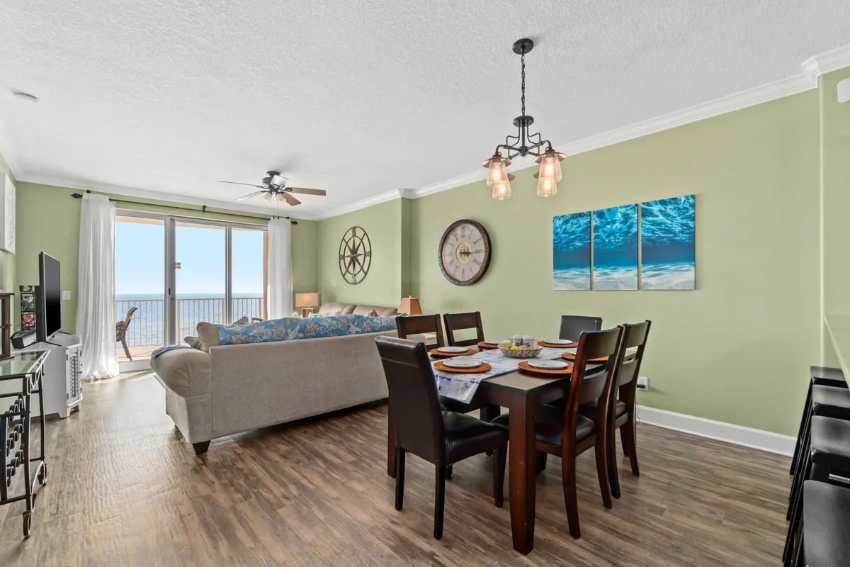 A welcoming open-plan living and dining area with large windows showcasing an ocean view. The dining table is set for a meal, and the comfortable sofa is accented with decorative pillows and colorful artwork on the walls. Natural light fills the space, highlighting the inviting and spacious layout.