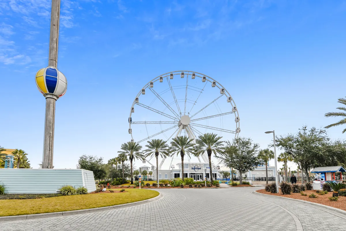 A festive Ferris wheel rises above palm trees and the entrance to the Sky Wheel attraction, with a clear blue sky overhead. The bustling scene hints at fun local activities just steps from the beach.