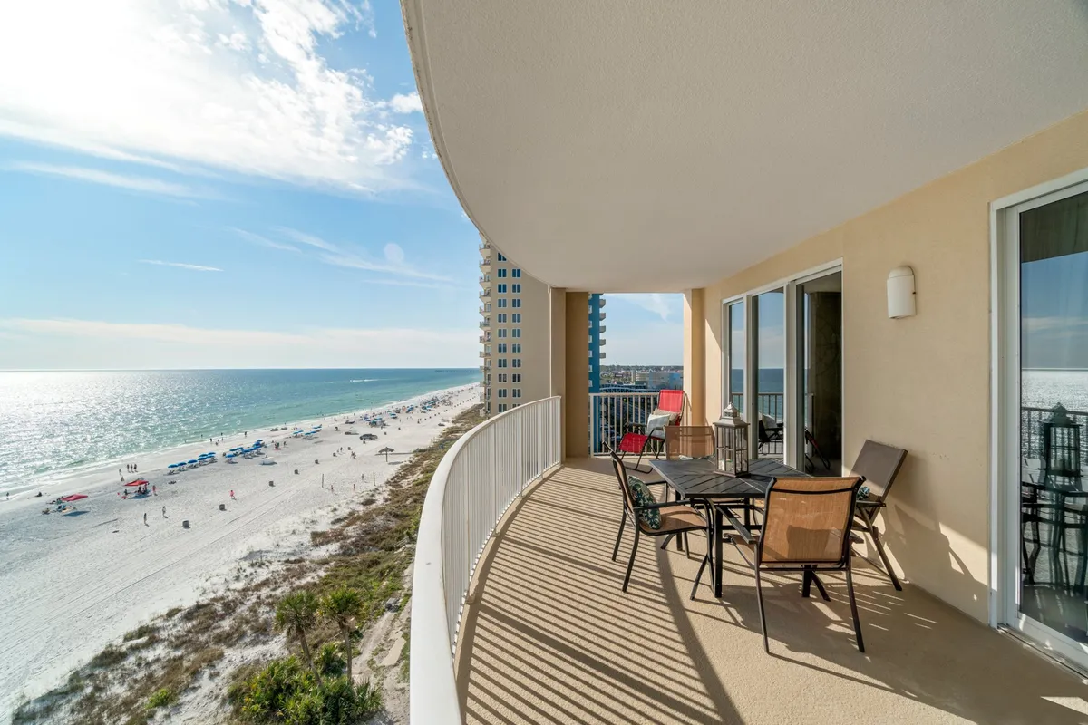 A serene view from a balcony overlooking the beach, featuring comfortable outdoor furniture set up for relaxation. The ocean waves are visible in the background and the setting sun casts a golden glow over the scene, suggesting a perfect spot for sunset drinks with friends.