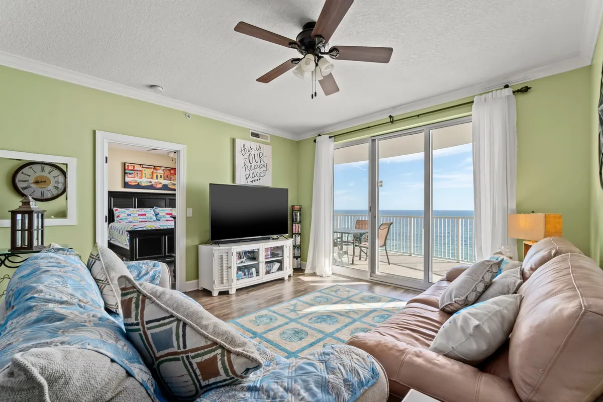 A bright, cheerful living room with comfortable seating (including a plush sofa), a flat screen television on a white stand, colorful decor accents, and large sliding glass doors opening to a balcony with an expansive ocean view. The room is filled with natural light and exudes a relaxed coastal vibe.