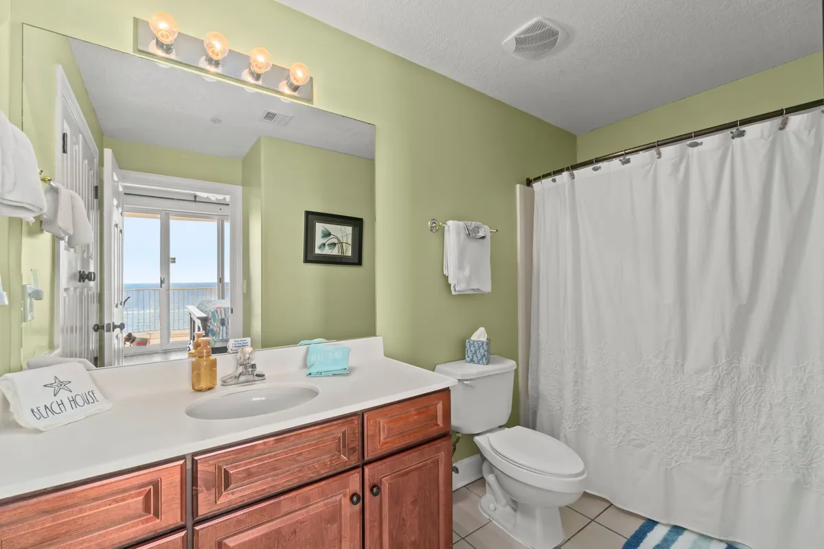 A fresh, modern bathroom featuring a large mirror reflecting an ocean view, a wooden vanity with a sink, a toilet with tissue box, and a shower with a white curtain. The space feels practical yet soothing, ideal for morning routines or unwinding after a day at the beach.
