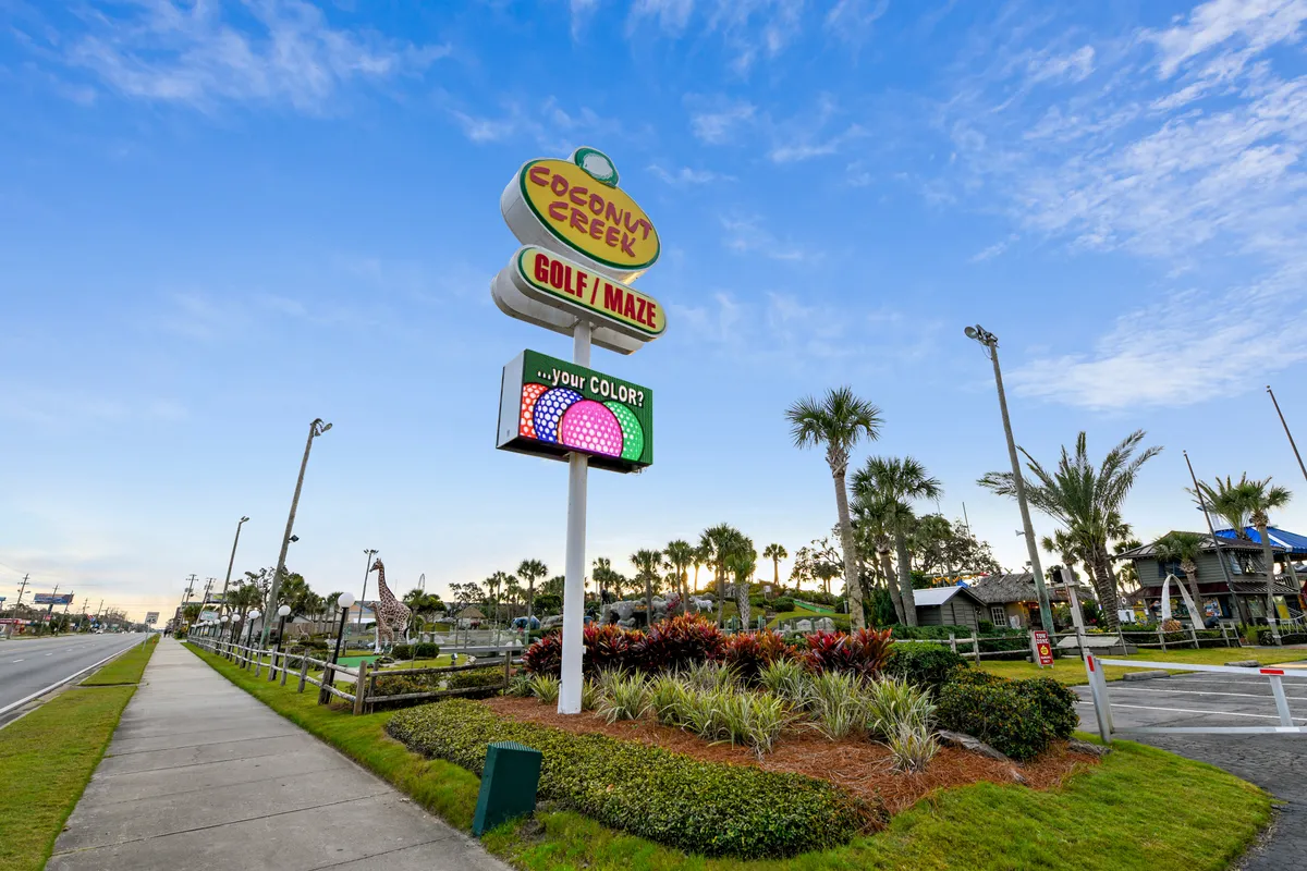 The entrance to Coconut Creek Golf & Maze features vibrant signage, a mini-golf course with playful decorations, palm trees, and inviting walkways. The area is lively and perfectly suited for group activities.